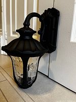 Front side angle view of the black aluminum wall light showing the scalloped top, curved arm, and textured glass.