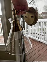 Side view photo of the wall sconce held by a hand, showing the clear conical glass shade with flared edges and brushed brass mounting plate and arm.