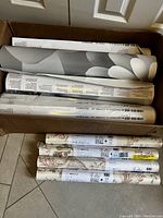 Box with three wall mural packs rolled, grayscale floral design on display. Beneath are four wallpaper rolls with ornate floral pattern labels.