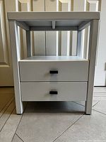 Front view of white wooden nightstand showing two drawers with black handles and bottom shelf.