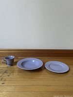 Set of three lavender ceramic items on wooden floor: one mug, one larger plate, one smaller plate showing scalloped ribbed edges.