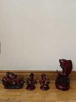 All four red resin figurines displayed on wooden floor against plain white wall, showing the full set.
