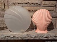 Photo showing both vases side by side against a stone background. Pink ceramic vase is shell-shaped with ribbed texture. Frosted glass vase has a matte translucent finish with shell-like ridges.