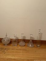 Full view of all five vintage crystal and glass decanters, including the covered pedestal bowl, arranged on floor against wall for display