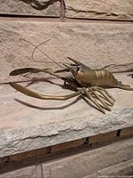 Metal lobster sculpture with detailed features placed on stone background, showing front and side views.
