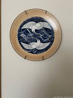 Front view of large round ceramic plate with twin cranes design in blue and white, textured beige border, and metal wall mount clips visible