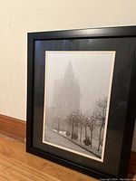 Framed black and white foggy landscape art print leaning against a wall on a hardwood floor, showing the print with lampposts and a tall building through fog.