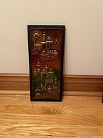 Full view of framed rectangular artwork showing intricate Middle Eastern cityscape design with metallic relief accents and a dark wood or black frame.