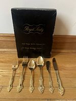 Box labeled Royal Sealy with 6 pieces of flatware (forks, spoons, knives) arranged in front showing decorative handles and gold plating.