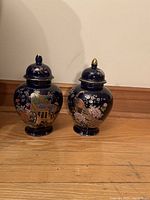 Two cobalt blue porcelain ginger jars with matching lids and intricate gold and floral designs shown standing on floor.