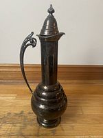 Front view of vintage silver plated water pitcher with conical lid on top, showing tall, slender design and ornate handle