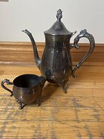 Silver plated teapot and creamer set on wooden floor, teapot closed with lid, showing ornate handle and feet.