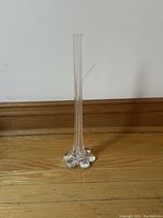 Full view of tall slender clear glass bud vase standing on wooden floor against white wall.