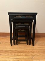 Four black lacquered nesting tables stacked inside one another showing detailed hand-painted designs on the edges.