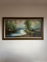 Front view of the large framed oil painting showing a river landscape with trees and foliage under a cloudy sky.