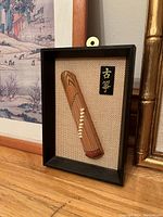 Miniature wooden guqin in black shadow box frame with textured beige backdrop and gold calligraphy plaque.