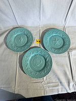 Three greenish-blue coastal themed ceramic plates from San Raphael Bordello displayed on a white cloth.