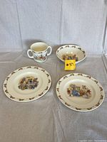 Five pieces of ceramic children's dishes featuring Beatrix Potter Bunnykins illustrations and bunny border pattern.