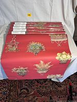 Six rolls of Thibaut wallpaper stacked horizontally showing decorative floral and planter motifs on red background.