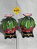 Pair of ceramic cherry and leaf designed tealight holders with black and gold bases and pink flower tops, shown standing side by side.