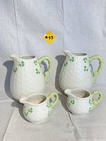 Photo showing a set of four ceramic pieces: two large pitchers and two smaller creamers or pitchers, white with basket weave texture and green shamrocks, bamboo-style handles.