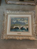 Framed painting of a French scene featuring a prominent stone bridge and architectural buildings in the background, ornate frame with decorative carvings and gold accents.