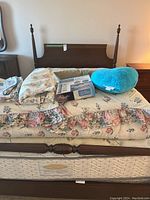 Vintage double wood bed frame visible with headboard, footboard, mattress, box spring, and bedding items including floral linens and blue heart pillow.