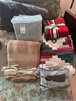 Overall lot of blankets, quilts, electric blanket, decorative cushion and throws arranged on a floral upholstered chair.