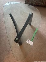 Top-down view of the coffee table showing the clear, irregular triangular glass top resting on a black base with three legs connected by silver metal fasteners.