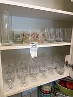 Shelves containing 6 short crystal stem wine glasses with leaf pattern and 3 tall clear drinking glasses along with other assorted glasses.