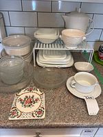 Photo of white Corelle dishes including square plates, bowls, a tea pot, glass bowls, and wire racks arranged on countertop with tiled backsplash
