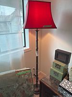 Full view of metal floor lamp showing red fabric shade and pole design situated near a window and beside decorative boxes.