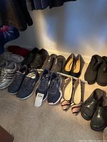 Multiple pairs of women's shoes arranged on carpet including athletic sneakers, flats, and boots, mostly dark colors, various styles and brands, some with tags, all needing cleaning.