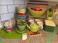 Full overview of assortment including stacked bowls, teapot, mugs, utensils, tea canister, artificial fruit bowl and placemats on kitchen counter