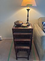 Stacked view of four dark wooden nesting tables with carved aprons, glass tops, two lidded boxes and table lamp on top.