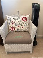 Front view of the white rattan chair with floral pillows and black tower fan next to it.