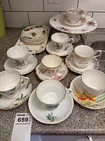 Full view of 8 teacups with matching saucers featuring floral and commemorative designs, stack of plates, and cake plate on tiered stand.