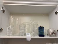 Full assortment of seven glass vases and containers inside a white cabinet shelf, showing variety of shapes, sizes and decorative rocks.
