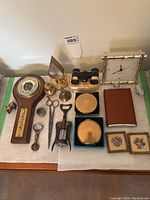 Full lot view showing wooden barometer, triangular clock, opera glasses, brass bells, bottle openers, scissors, corkscrew, two compact cases, brown case, framed floral artwork