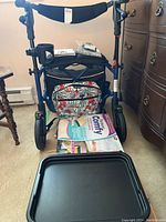 Blue three-wheeled Human Care Rollator Nexus 3 walker with floral storage bag, lap tray in front, combination seat cushion, and manual on walker platform.