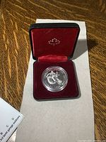 Silver dollar coin sealed in clear capsule displayed in red velvet-lined black case with maple leaf emblem inside lid.