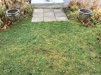 Wide shot of two concrete garden urns positioned symmetrically on a lawn next to a pathway, showing overall scale and placement.
