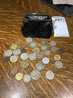 Photo showing the full lot including a black hexagonal pattern change purse and a large assortment of vintage foreign coins laid out on a wooden surface.