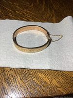 Top view of hinged oval gold-tone bangle on white background, showing clasp and safety chain