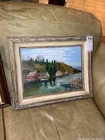 Full view of signed oil painting in frame showing a lake or river scene with houses, trees, dock and boats.