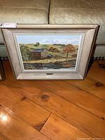Framed landscape painting showing a rustic barn, trees and hills under a partly cloudy sky, placed on a wooden floor in front of a leather sofa.