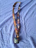 Full view of the vintage brass Swiss cow bell with a hand painted Edelweiss flower, hanging from a long floral fabric strap, placed on a blue cloth background.