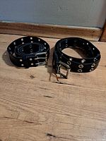 Two black punk belts with double metal grommets and metal buckles laid on wooden floor.