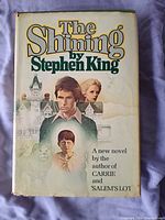 Front cover of the book The Shining by Stephen King with illustrated dust jacket showing characters and buildings.