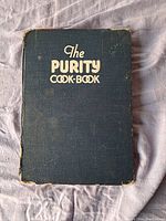 Front cover of The Purity Cook-Book hardcover cookbook, showing black textured cover with gold embossed title in worn condition.